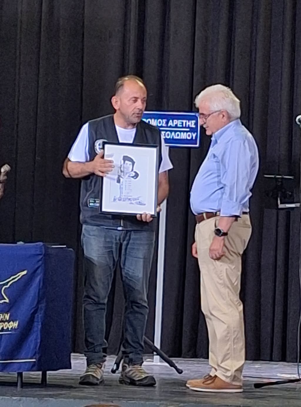 Man in a vest presents a framed drawing to an older man on stage; both wear casual attire, with a blue sign and black curtain backdrop behind them