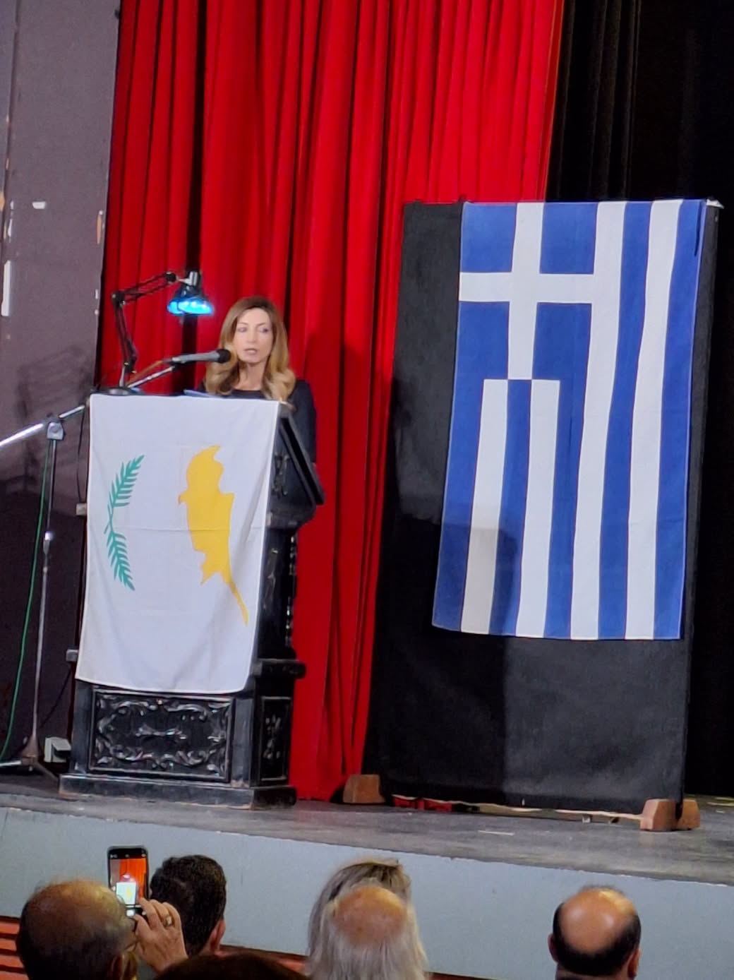 Woman speaking at a podium on a stage with red curtains and a Greek flag backdrop.