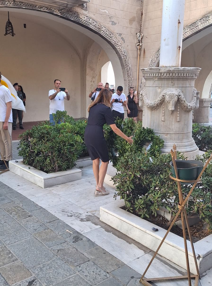 Woman in a black dress tending a shrub bed near a decorative stone column while several people photograph the scene in a historic courtyard.