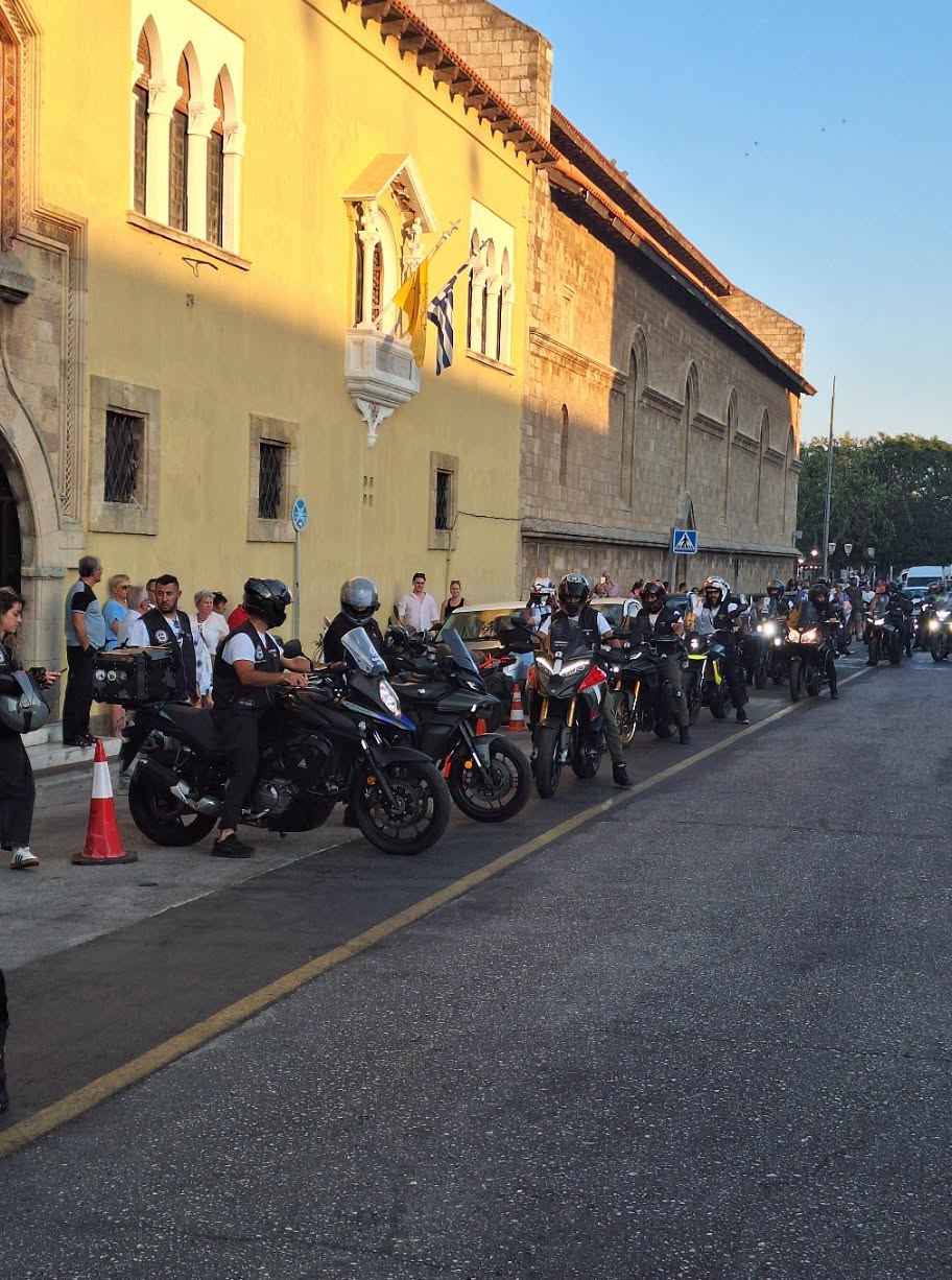 Row of parked motorcycles along a sunlit street beside a yellow historic building with flags and onlookers.