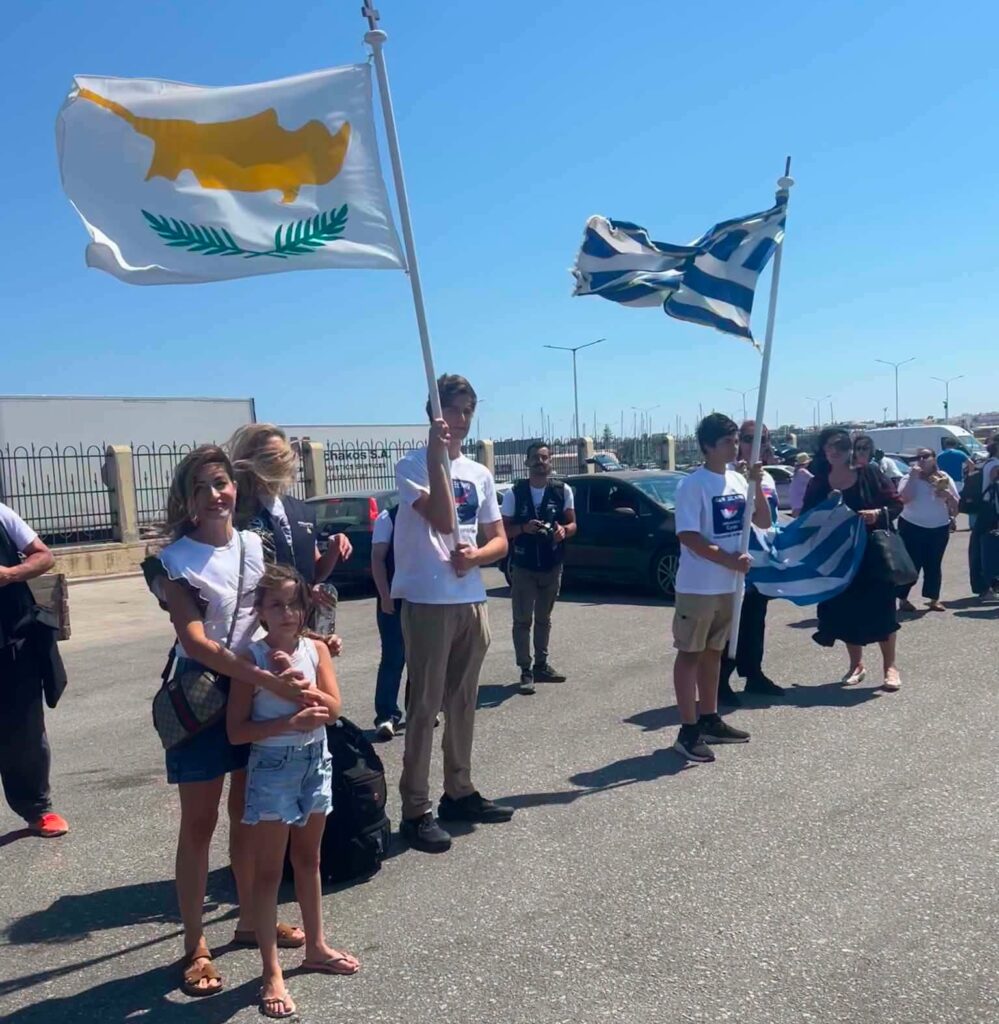 People in a parade holding two large flags, one white with an orange map and olive branches and a blue‑and‑white Greek flag, on a sunny street.