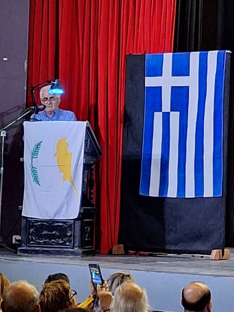Man speaks at a podium on a stage with red curtains, microphone nearby, audience watching in front; Greek flag displayed beside the stage.