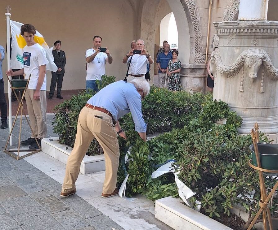 Man in a blue shirt and tan pants leans over a hedge planter, arranging a wreath during a ceremony as people photograph the scene in the background.