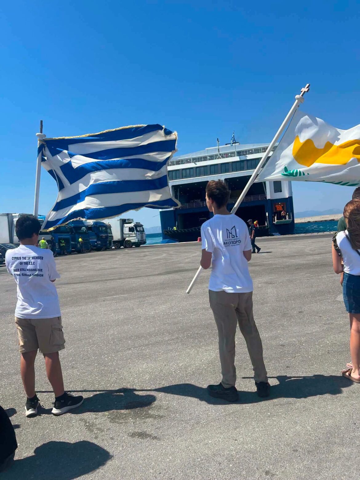 Port scene with a blue-and-white Greek flag and a white flag with a yellow emblem in front of a large ferry on a sunny day