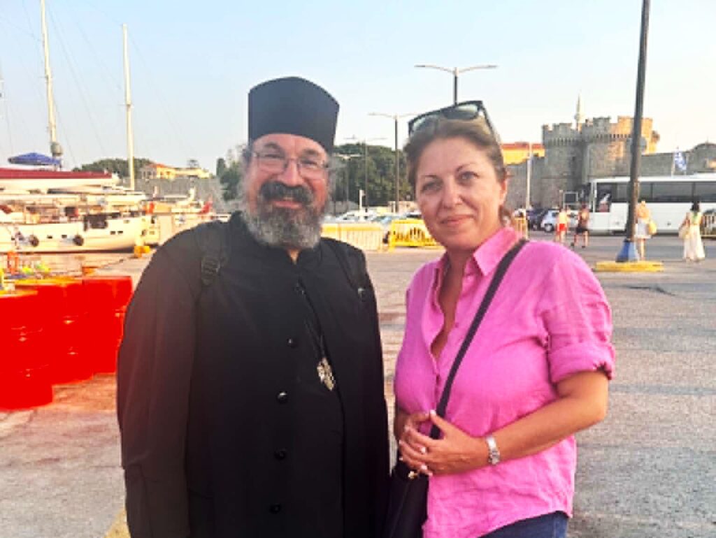 Man in black clerical robes and a woman in pink stand together at a marina, smiling.