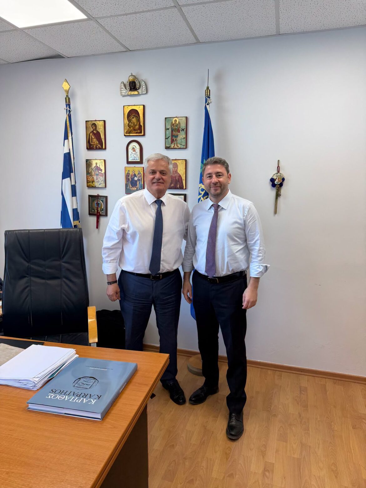Two men in dress shirts and ties stand together in an office, with flags and icon wall decor behind them.