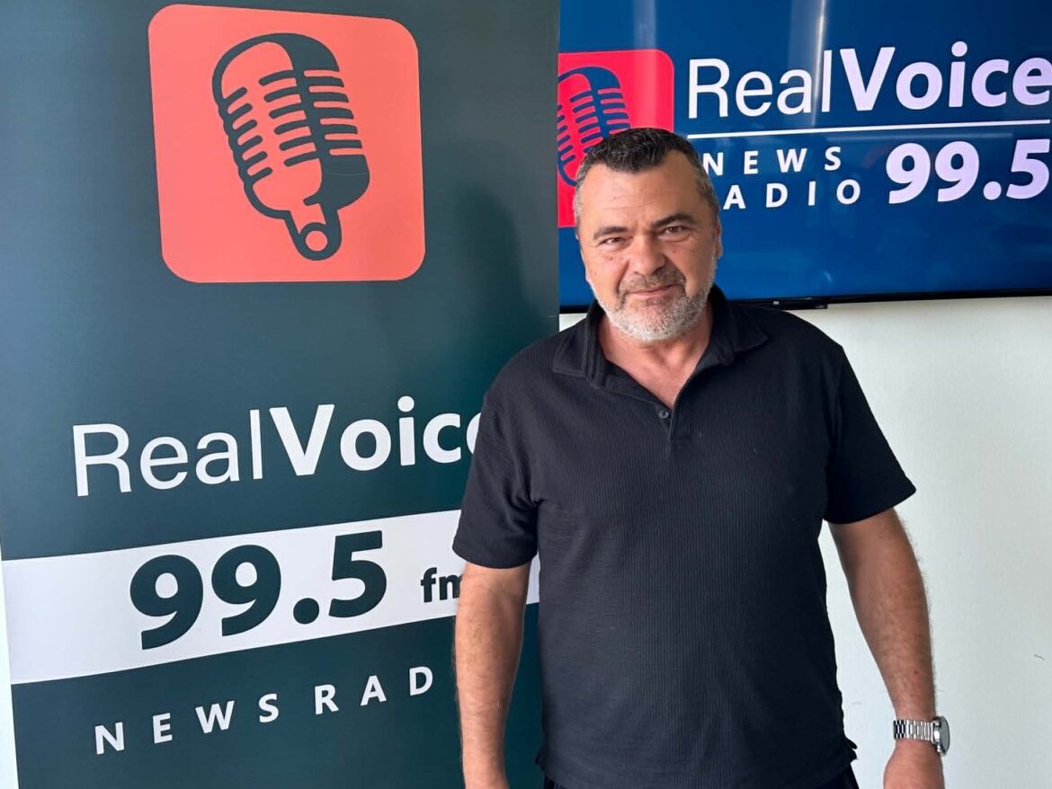 Man posing beside a RealVoice news radio banner with a red microphone logo and '99.5 fm' visible.