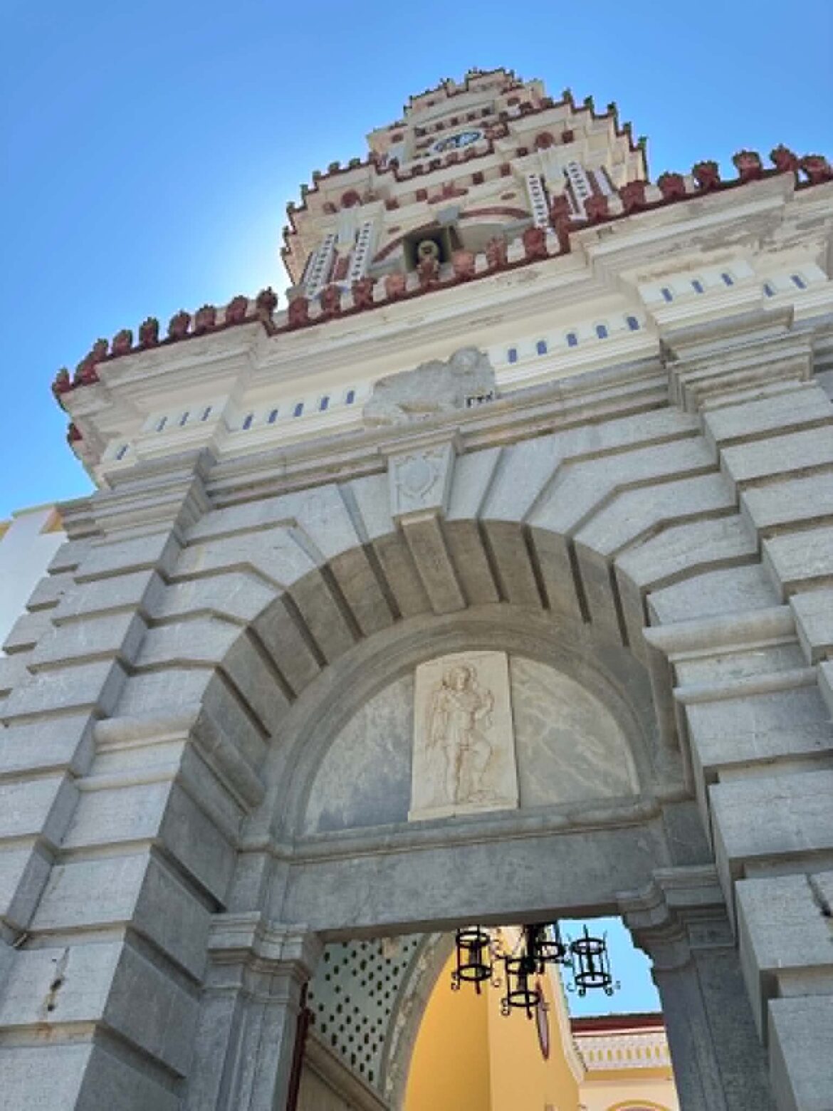 Stone arched gateway in gray stone with decorative stepped top, opening to a bright yellow courtyard beyond against a blue sky.