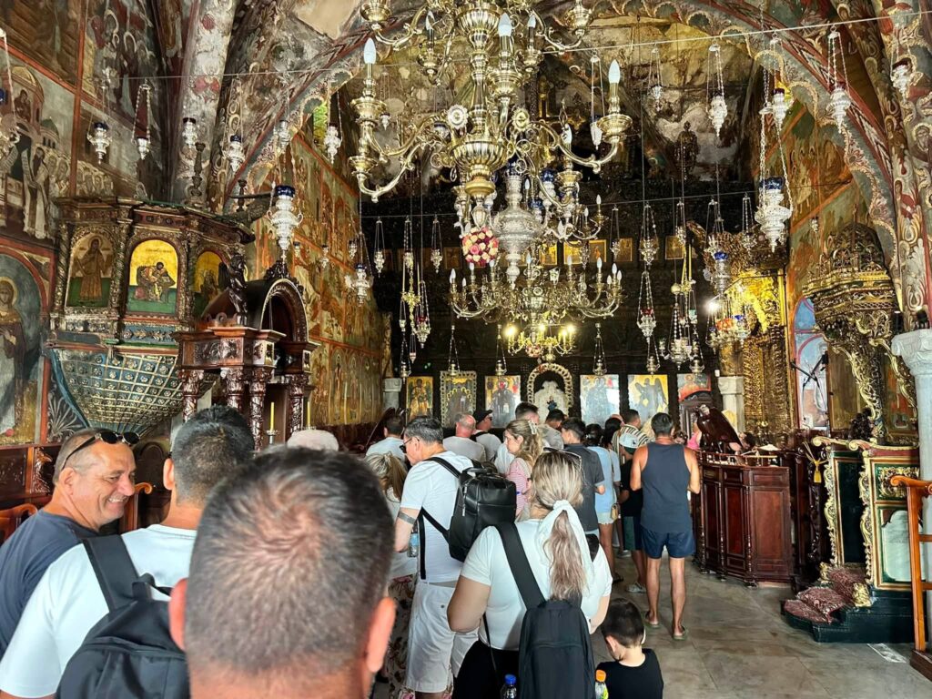 Crowded interior of an ornate Orthodox church with gilded chandeliers, murals, and icons along the walls.