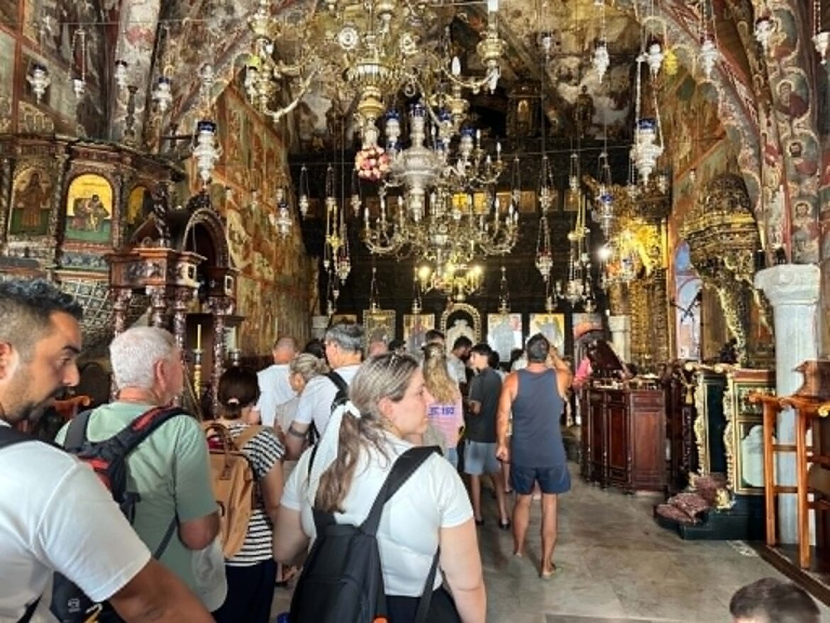 Crowded ornate church interior with gold altarpieces, icons, and chandeliers as visitors wait in a line.