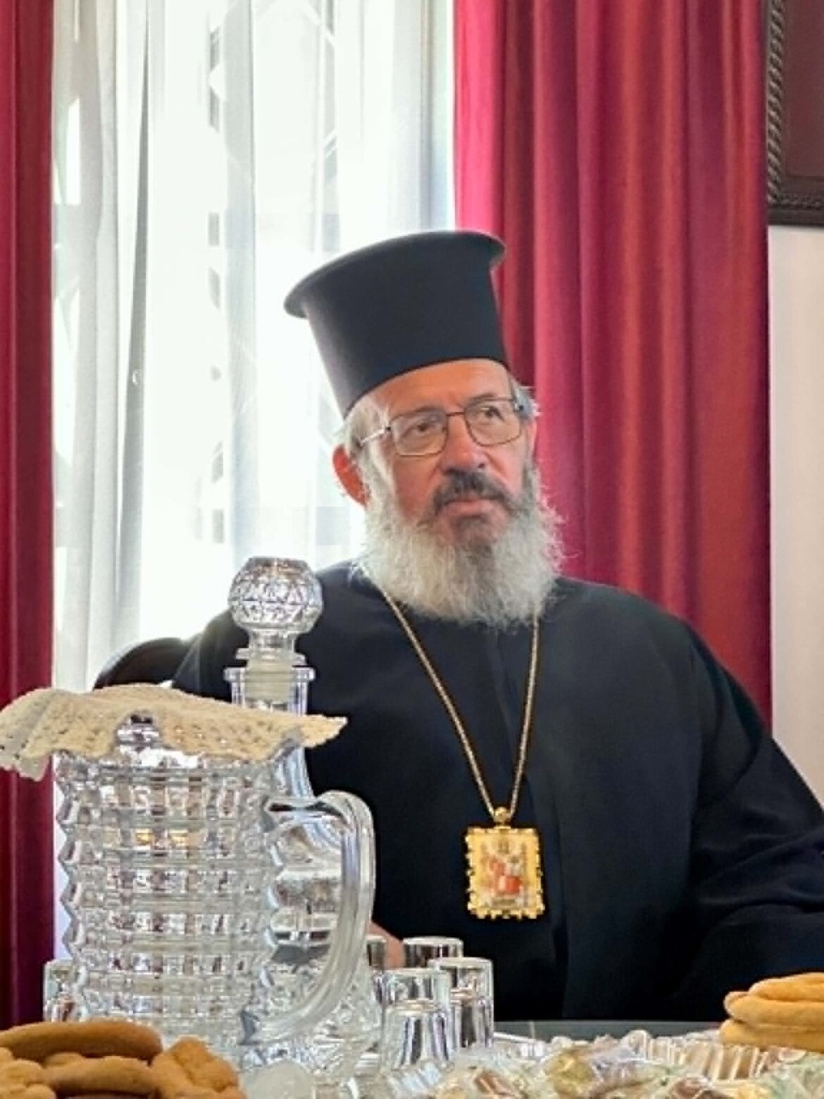 Orthodox priest in black robes and tall hat, wearing a gold cross, seated at a table with ornate silver dishes and pastries against red curtains.