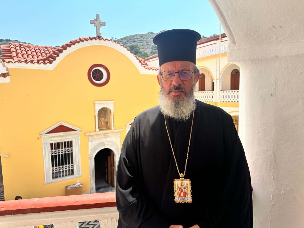Orthodox priest in black robes and a tall hat stands in a sunlit courtyard beside a bright yellow church with red-tiled roof and a cross on top.