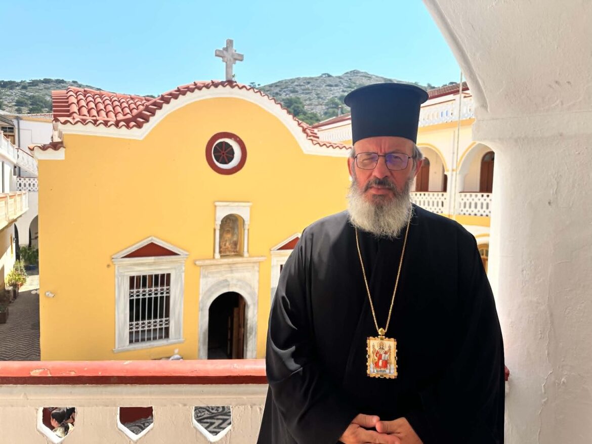 Orthodox priest in black robes and a tall hat stands in front of a bright yellow church with a red-tiled roof and cross on top.