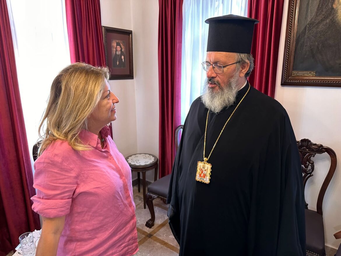 Woman in a pink shirt speaks with an Orthodox priest wearing a black robe and tall hat in a room with red curtains and framed art.