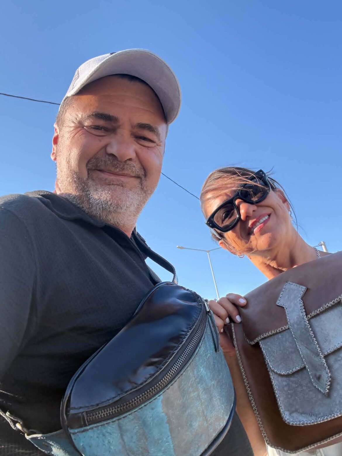 A man and woman taking a selfie outdoors against a clear blue sky, smiling at the camera, with a gray cap and sunglasses visible.