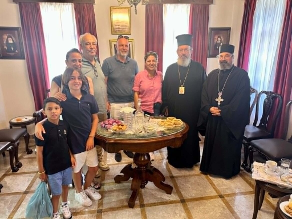 A mixed group posing for a photo: two Orthodox priests in black robes stand with others in a room, gathered around a table of pastries.