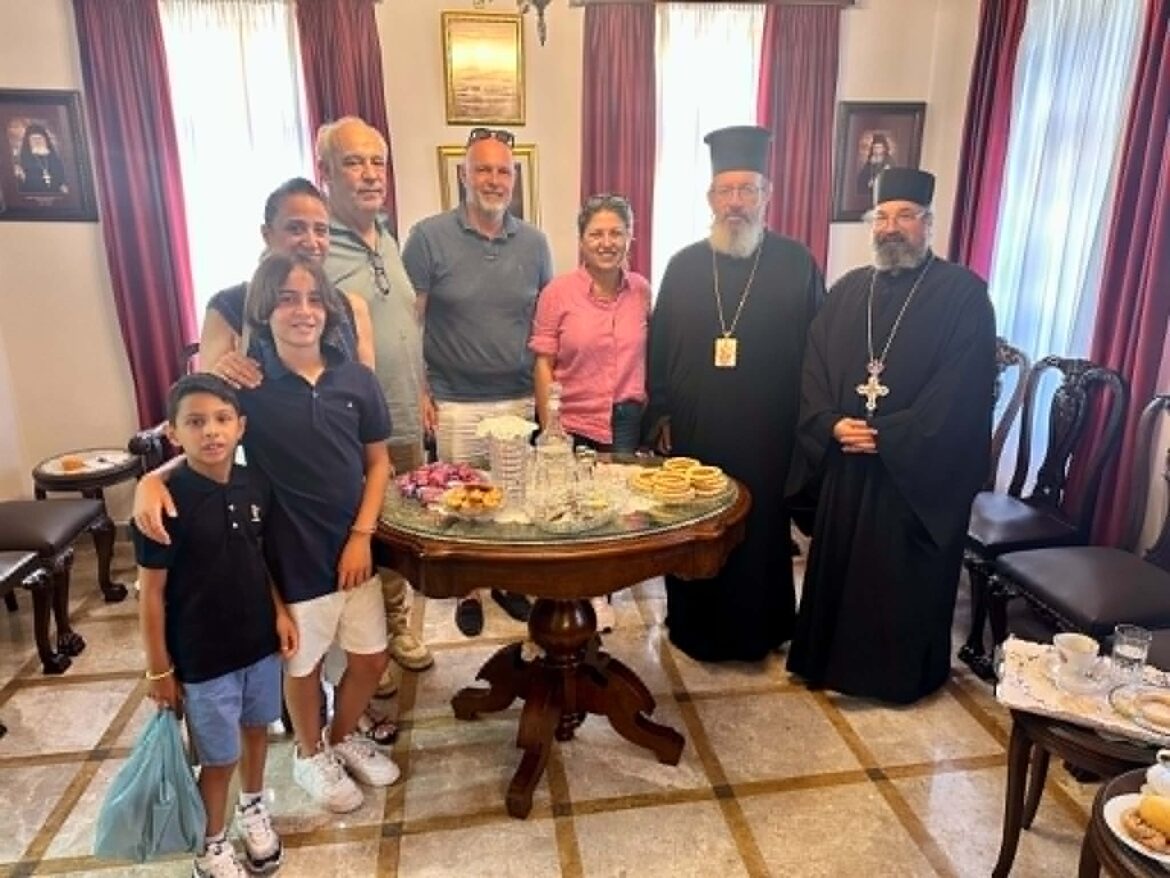Group of eight people posing behind a table of pastries in a formal room; two Orthodox priests in black robes stand on the right, with family members smiling.