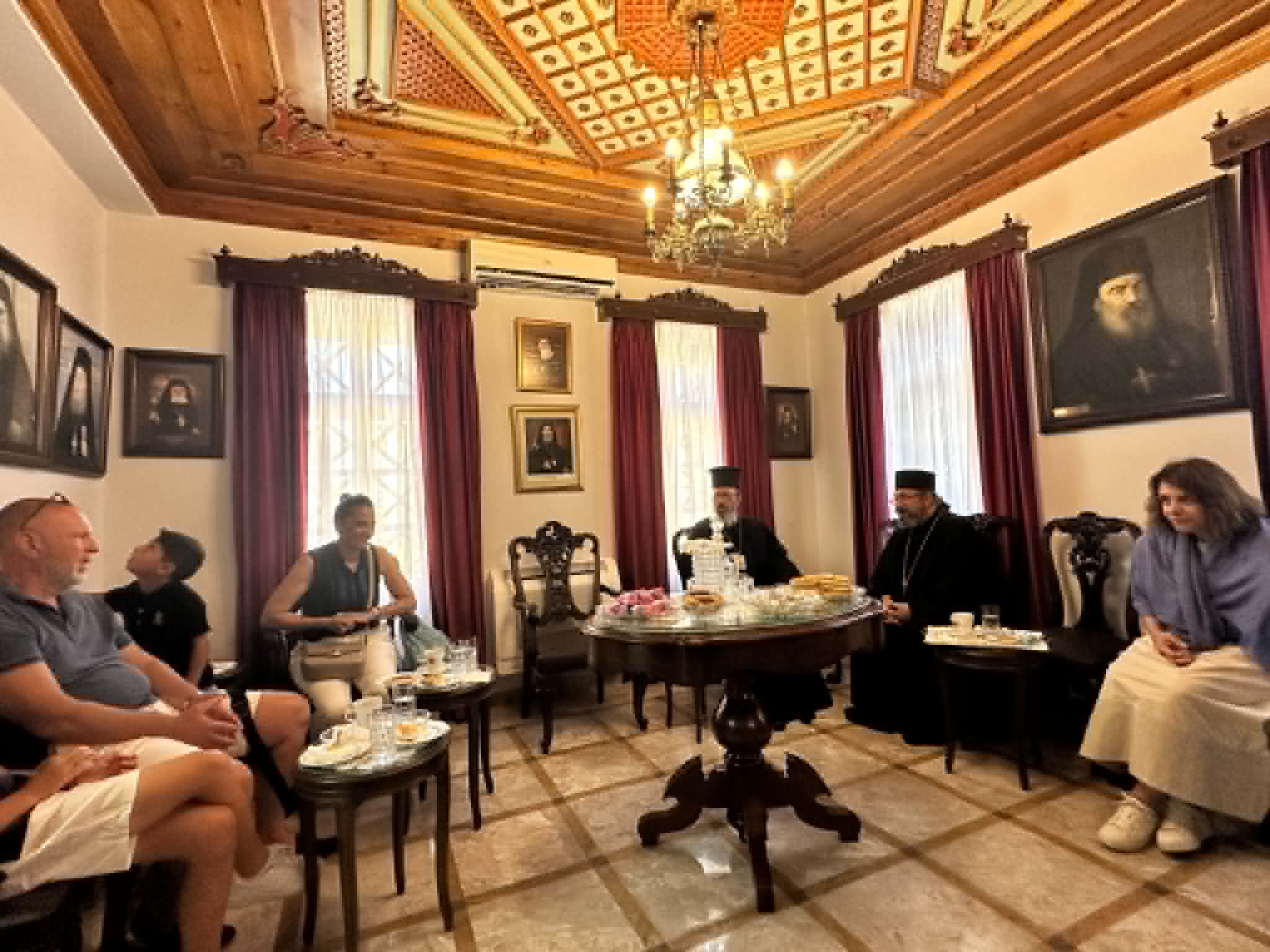 Formal dining room scene with a chandelier and ornate wood-paneled ceiling; people seated around small tables, chatting over tea and pastries, portraits on the walls framing the gathering.