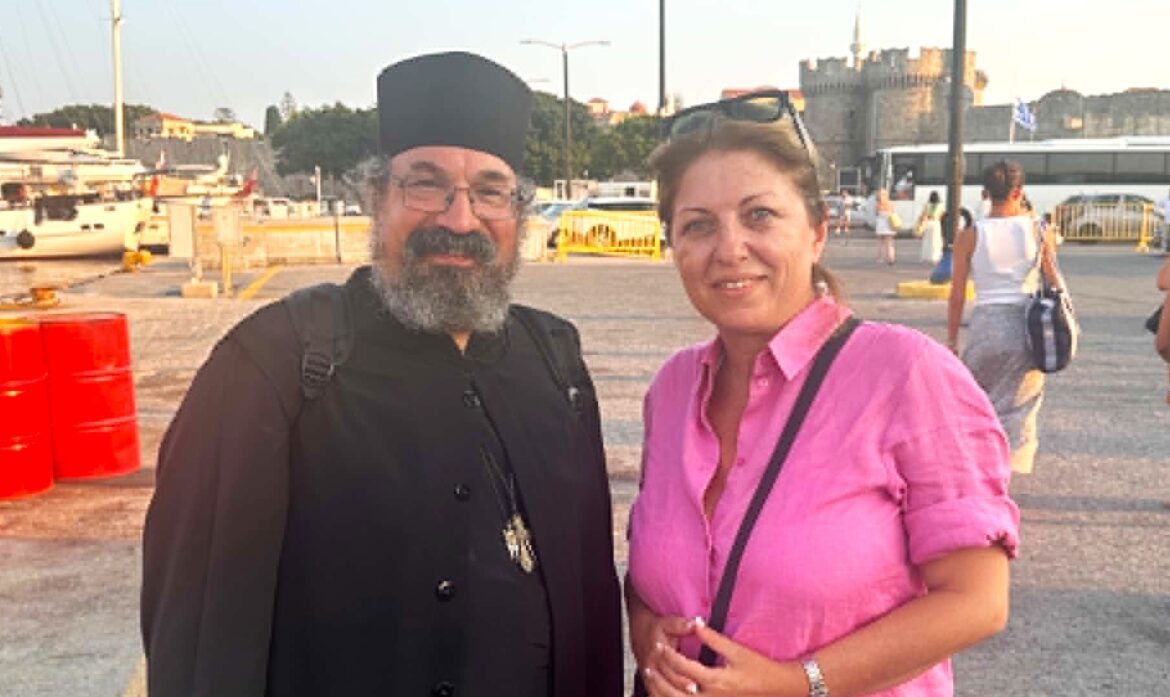 Two adults posing together outdoors: a bearded man in black clerical attire and a woman in a pink shirt smiling at the camera.