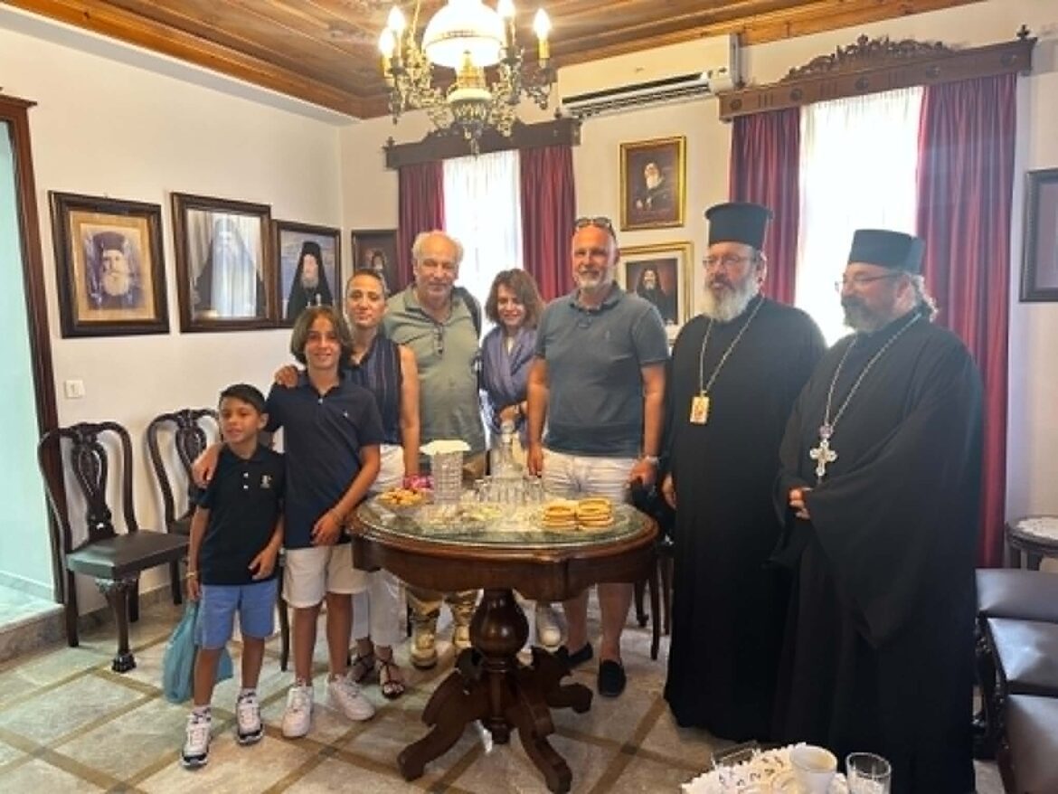 Group of people posing around a round table with snacks in a formal room, with portraits on the wall and a chandelier above; two clergy in black robes stand on the right.