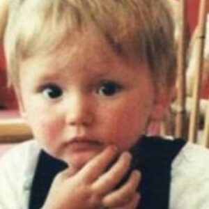 Toddler with blond hair looking at the camera, hand resting on chin in a thoughtful pose